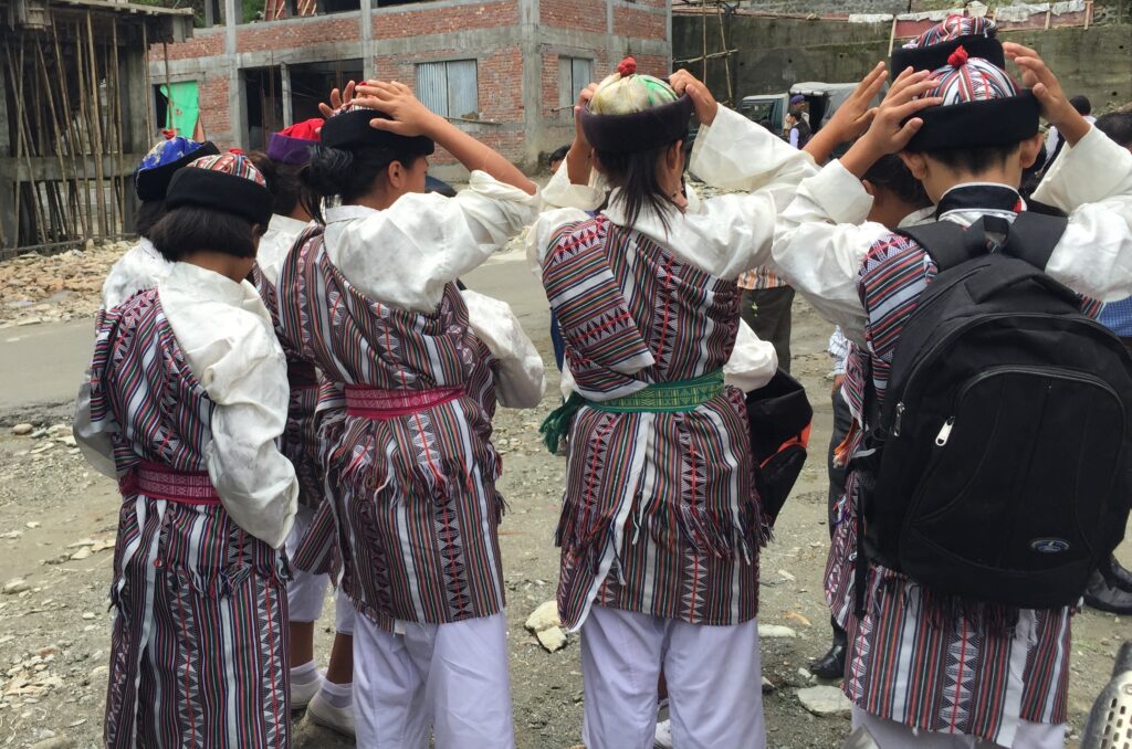 From behind, a photograph features a group of children wearing patterned hats and clothing—some with black backpacks—standing in a village square. All but a few children in the back of the group have both hands on their heads.
