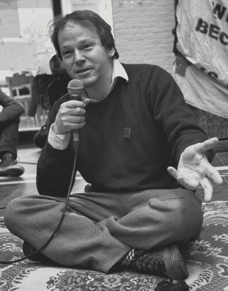 A man sits cross-legged on a patterned rug, holding a microphone and gesturing while speaking.